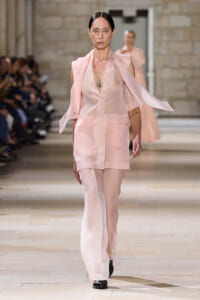 Model walking a fashion runway in a pale pink sheer suit with cape-like sleeves, tattoo visible on her chest, audience on both sides.