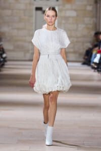 Model on a runway wearing a white textured short dress with a gathered bubble skirt, slim belt, and white ankle boots.