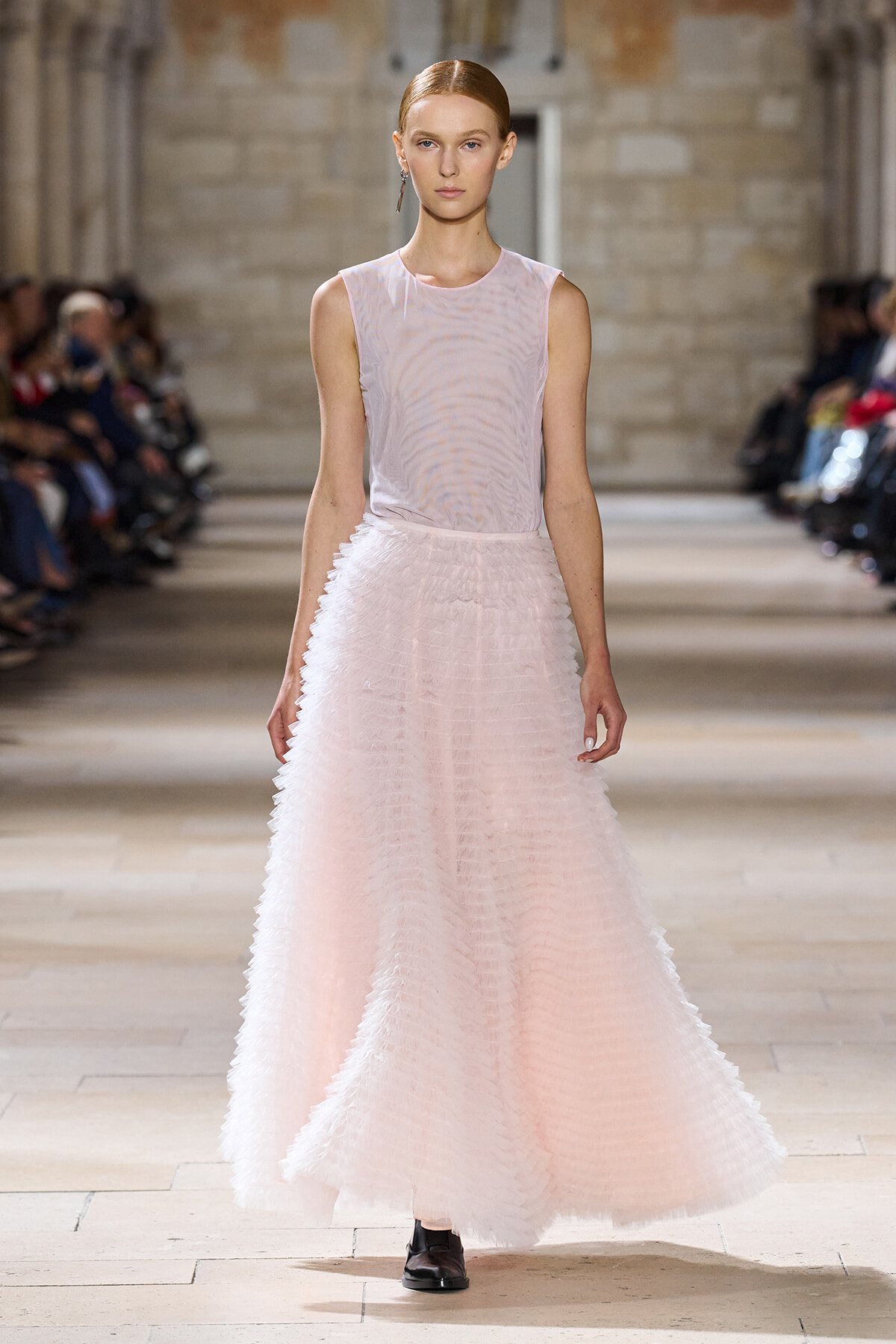 Model walks the runway in a soft pink sleeveless top and a full, textured pale pink skirt; audience on either side in a stone-walled venue.