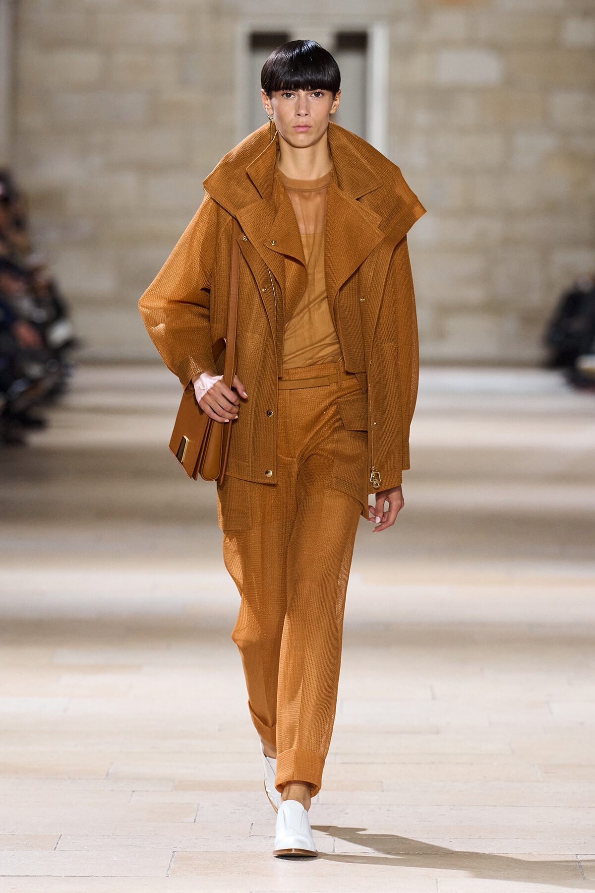 Model walks a fashion show wearing a monochrome camel-brown outfit: oversized coat over a matching suit, carrying a brown handbag, and white shoes.