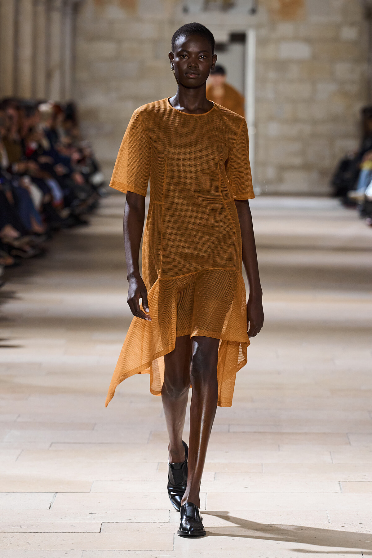 Model walking runway in orange mesh tunic dress with asymmetrical hem, black loafers, audience blurred in background