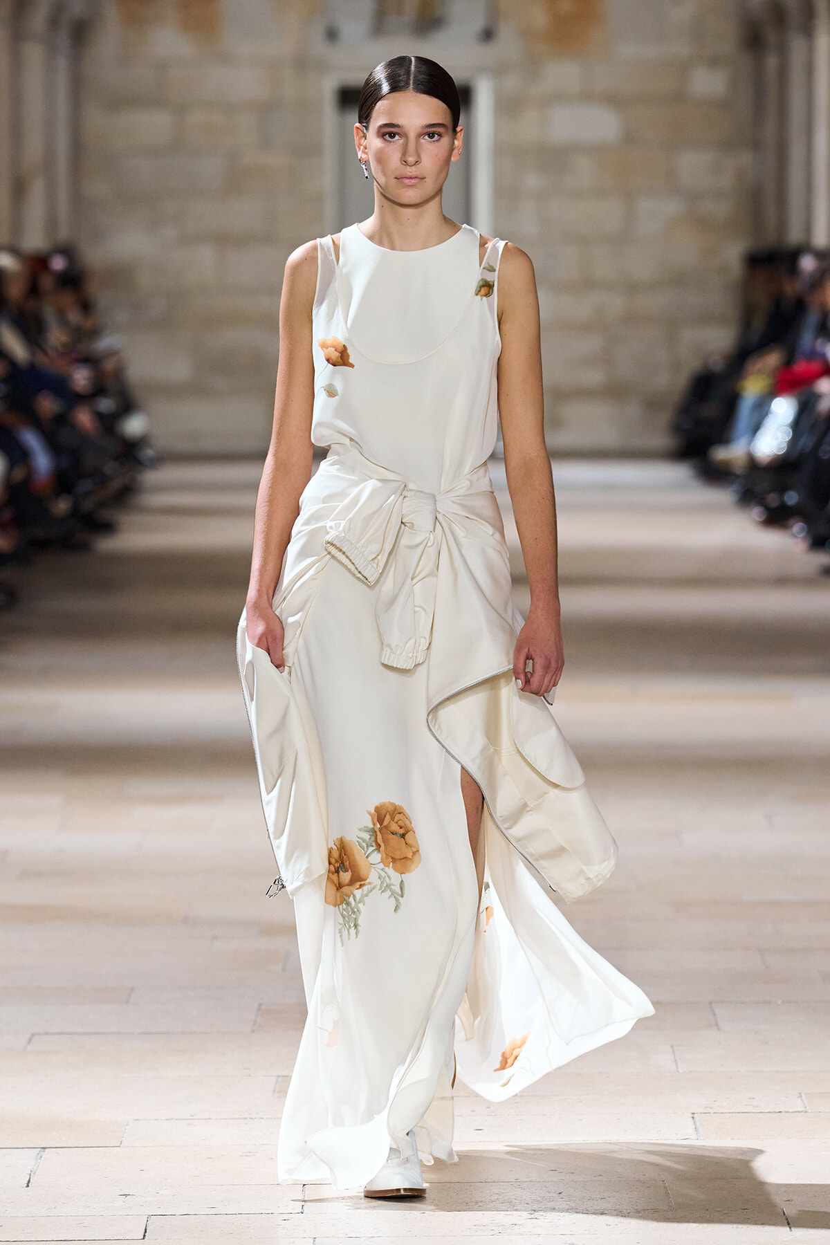 Model walking down a runway in a white sleeveless gown with orange floral embroidery and a tied waist detail.