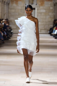 Female model walks the runway in a sculptural white one-shoulder dress with cascading ruffles.