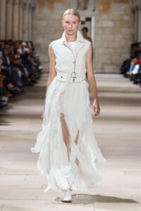 Model walks a runway wearing a white sleeveless trench dress with a belted waist and flowing asymmetric skirt pieces.