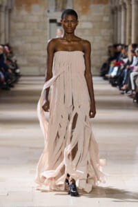 Model walks a runway in a strapless, pale pink, pleated gown with ribbon-like strips flowing as she strides forward.