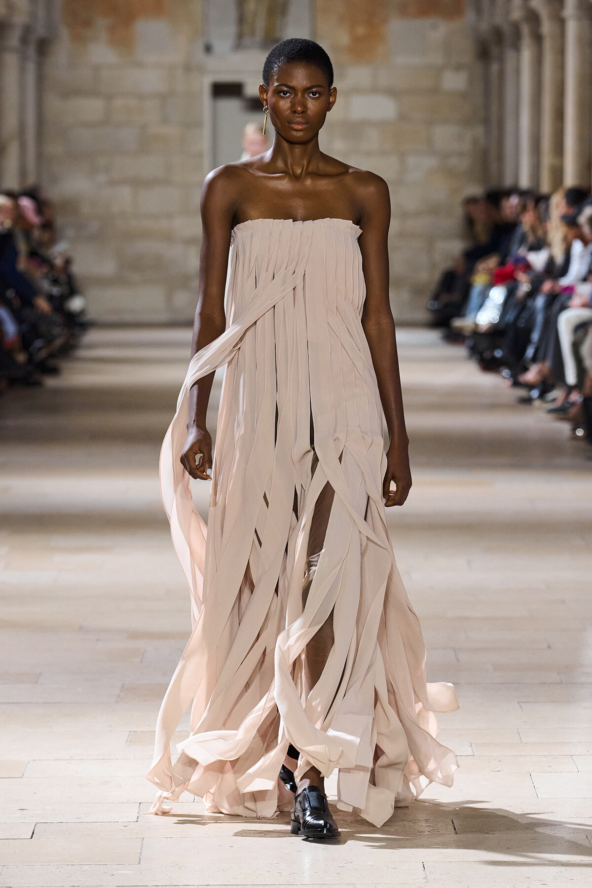 Model walks a runway in a strapless, pale pink, pleated gown with ribbon-like strips flowing as she strides forward.