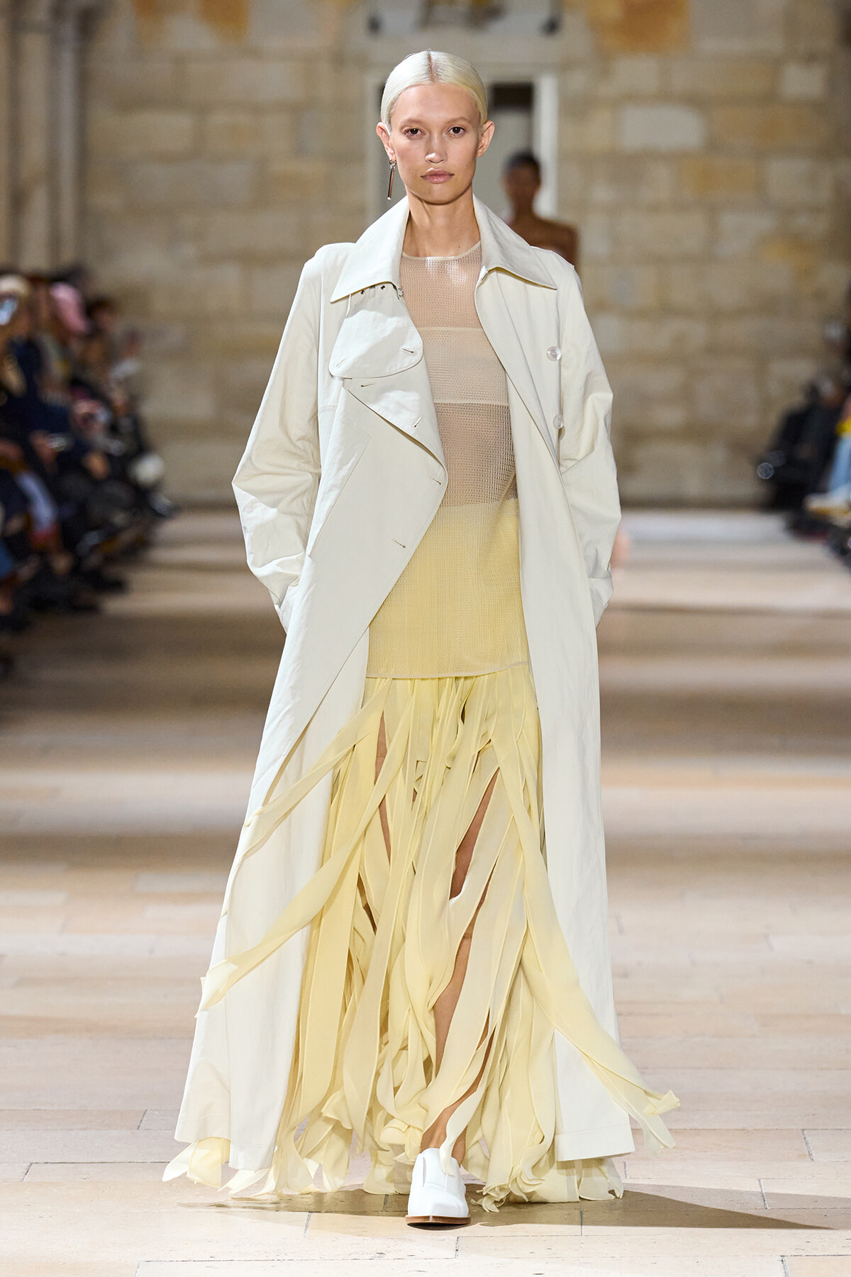 Model walks a runway wearing a long pale trench over a sheer beige knit top and a pale yellow fringe skirt, with white shoes.
