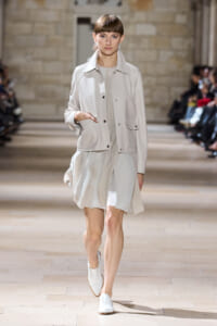 Model walking a fashion runway in a light beige jacket over a pale, flowing dress and white shoes in a stone-walled venue.