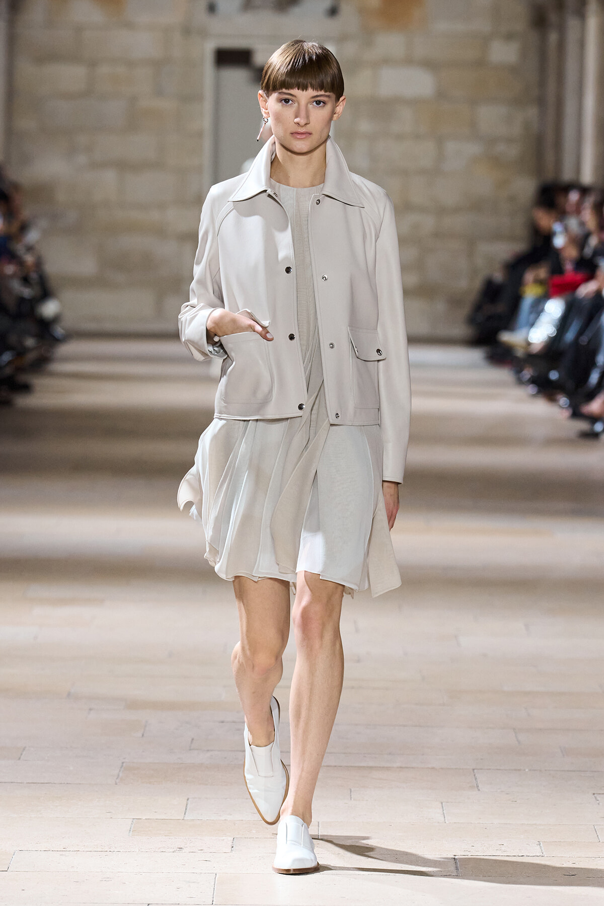 Model walking a fashion runway in a light beige jacket over a pale, flowing dress and white shoes in a stone-walled venue.