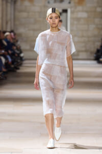 Model walks the runway in a sheer white two-piece outfit with geometric patch patterns.