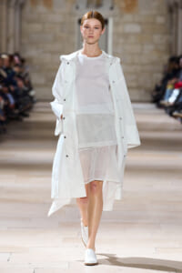 Model walks a long white coat over a sheer white dress on a runway left-to-right catwalk.