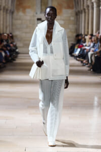 Model on a runway wearing an all-white, mesh suit with an oversized blazer, sheer top, and wide-leg pants, carrying a white clutch bag.