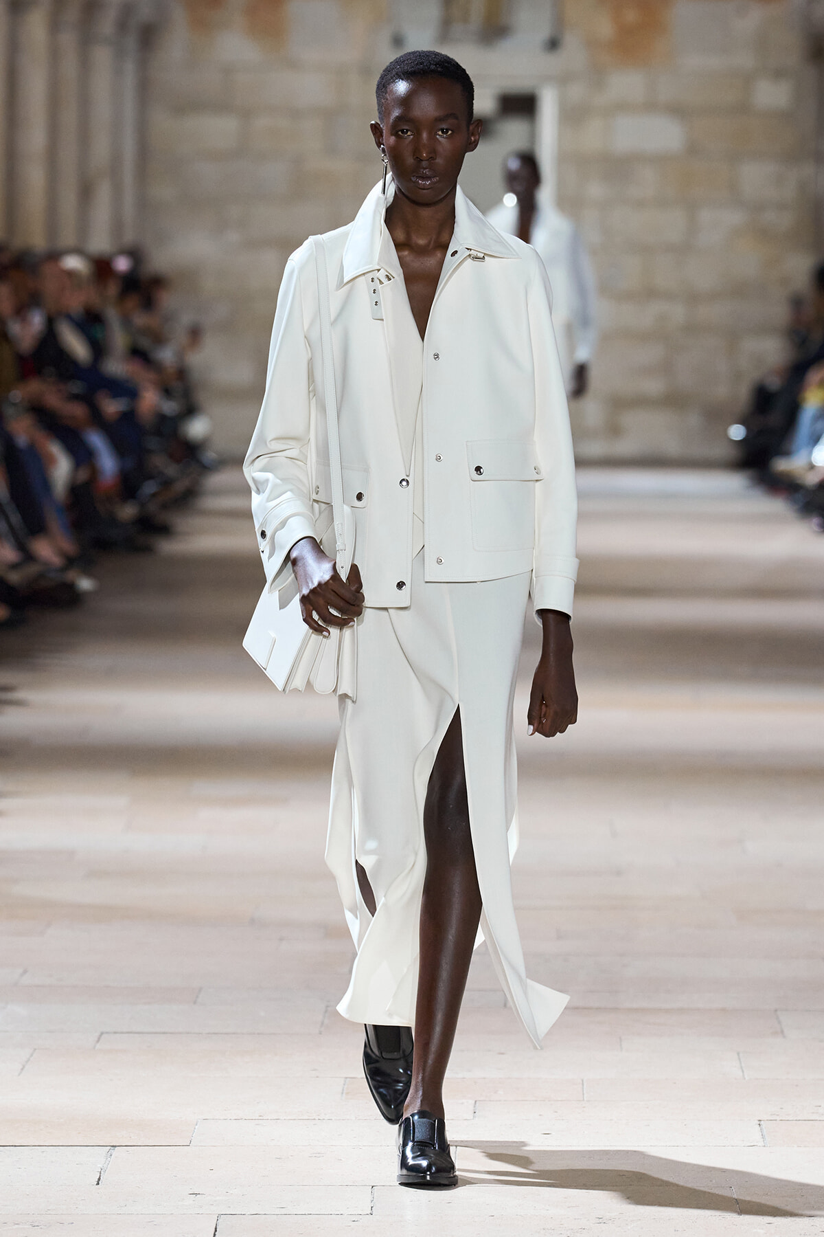 Female model walking a fashion runway in an all-white, tailored suit with a long coat and matching skirt with a front slit, carrying a white handbag.