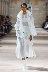 Female model in an all-white sheer outfit with a white jacket walking a fashion show runway