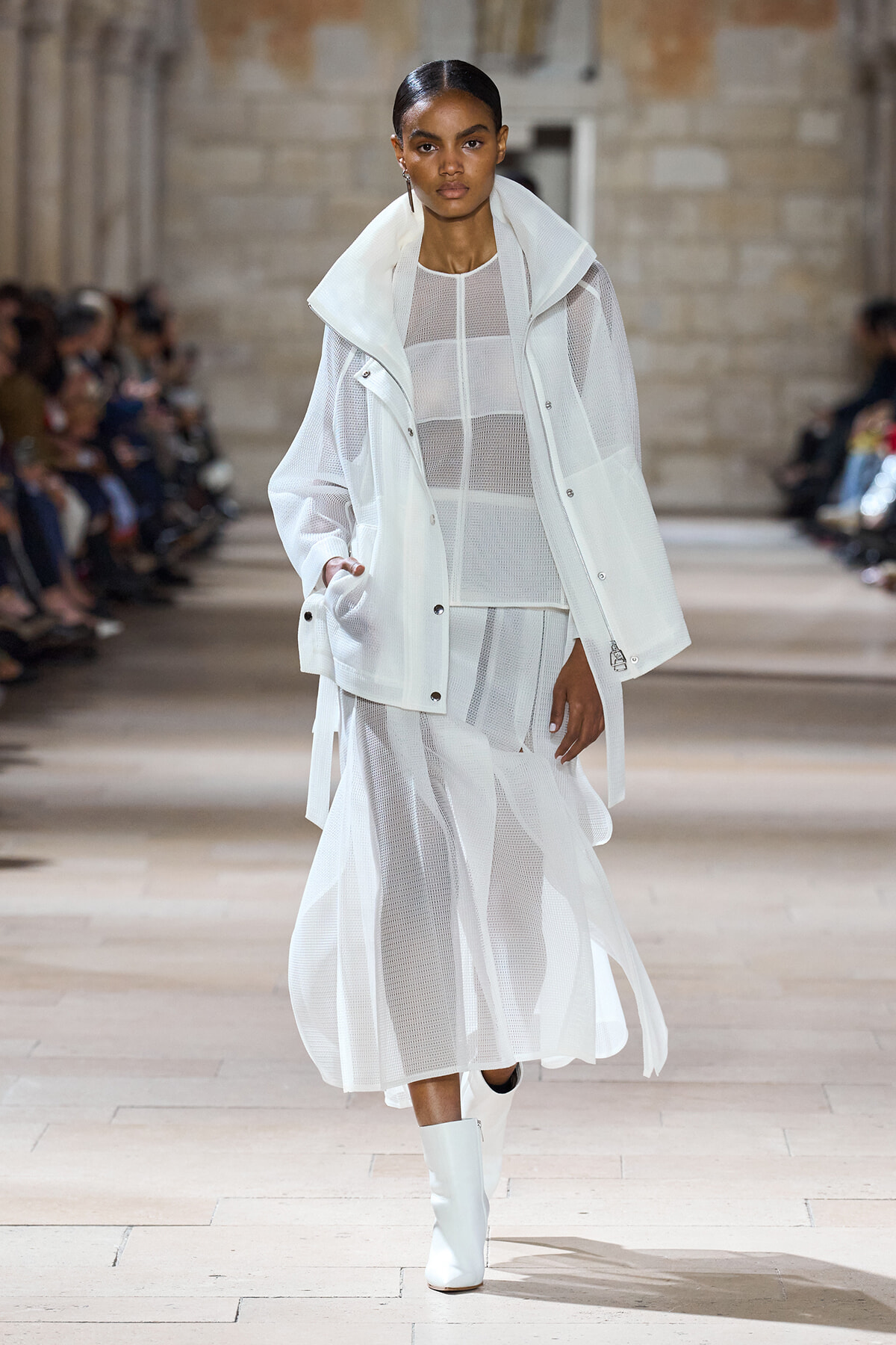 Female model in an all-white sheer outfit with a white jacket walking a fashion show runway