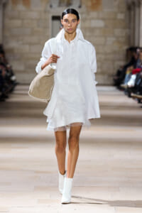 Model on a runway wearing an all-white ensemble: oversized white shirt, layered sheer skirt, white ankle boots, and a large beige shoulder bag.