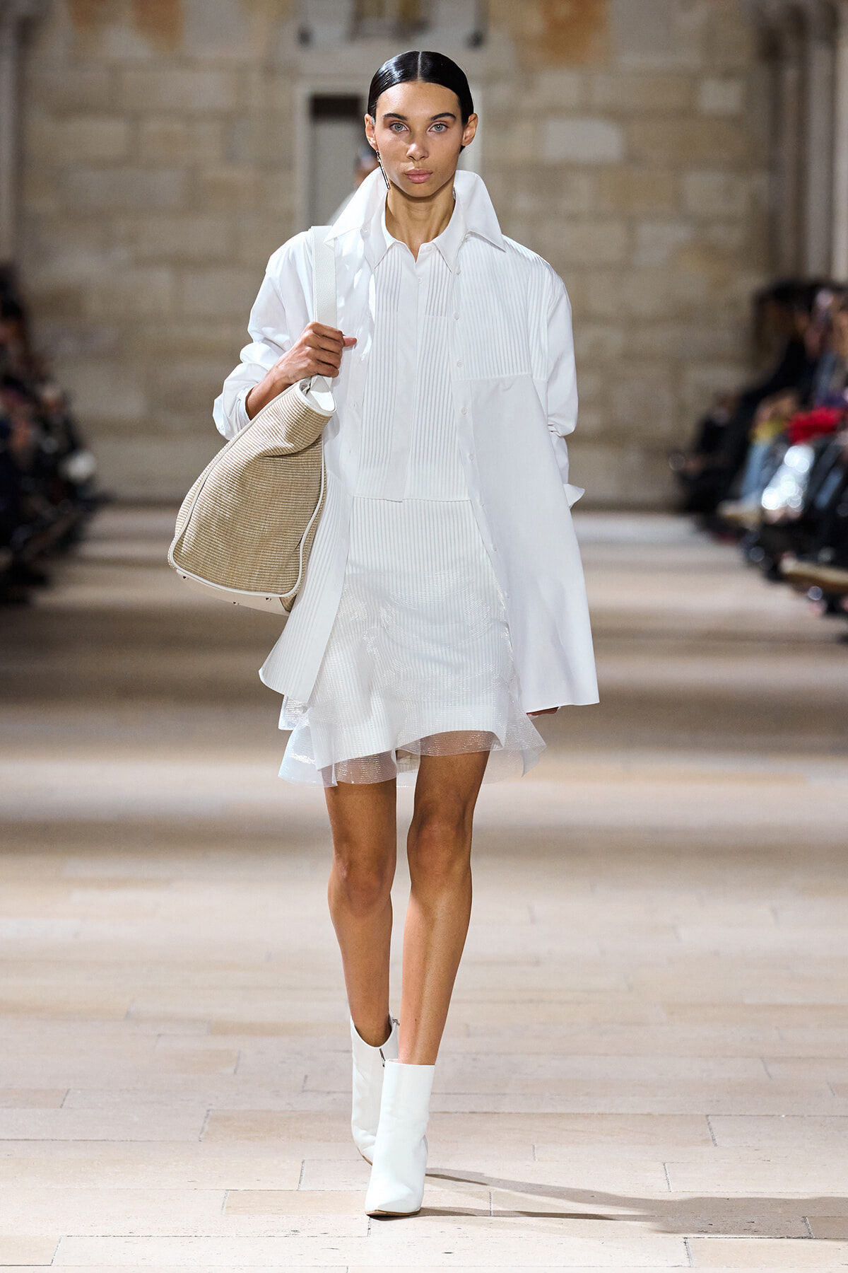 Model on a runway wearing an all-white ensemble: oversized white shirt, layered sheer skirt, white ankle boots, and a large beige shoulder bag.