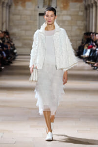 Model walking a runway in an all-white outfit: textured fluffy jacket, sheer top, layered midi skirt, white shoes, and a white handbag.