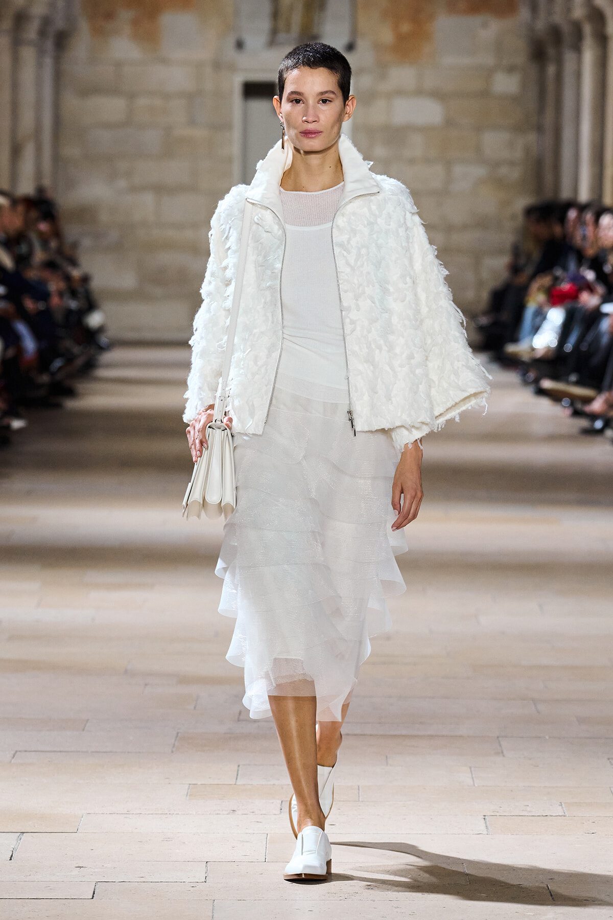 Model walking a runway in an all-white outfit: textured fluffy jacket, sheer top, layered midi skirt, white shoes, and a white handbag.