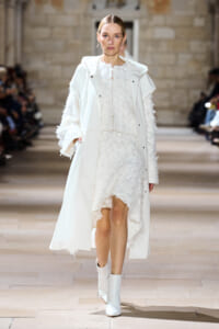 Model walks a runway in an all-white outfit: a long white coat over a textured white dress with lace details and white ankle boots.