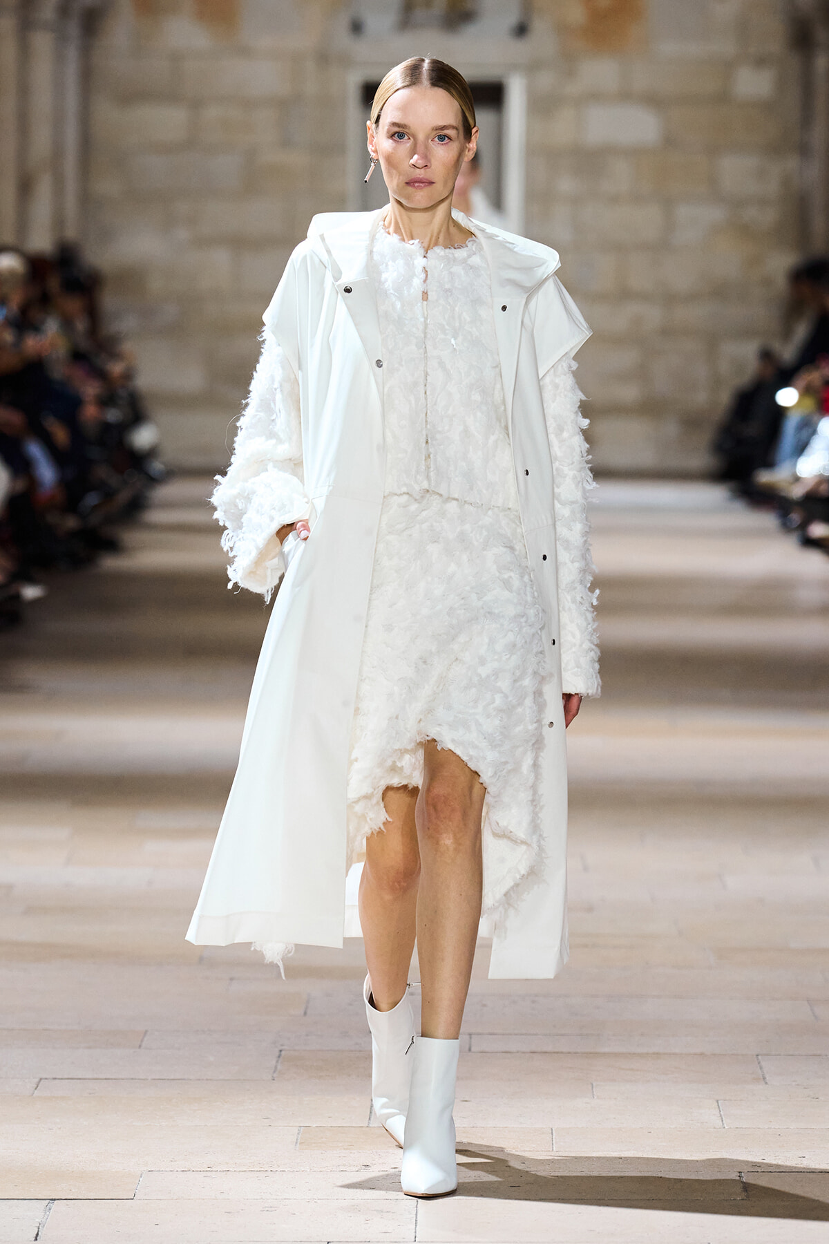 Model walks a runway in an all-white outfit: a long white coat over a textured white dress with lace details and white ankle boots.