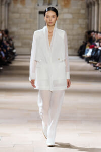 Model wearing an all-white layered blazer and wide-leg pants walks a fashion runway, sheer sleeves and structured detailing visible.