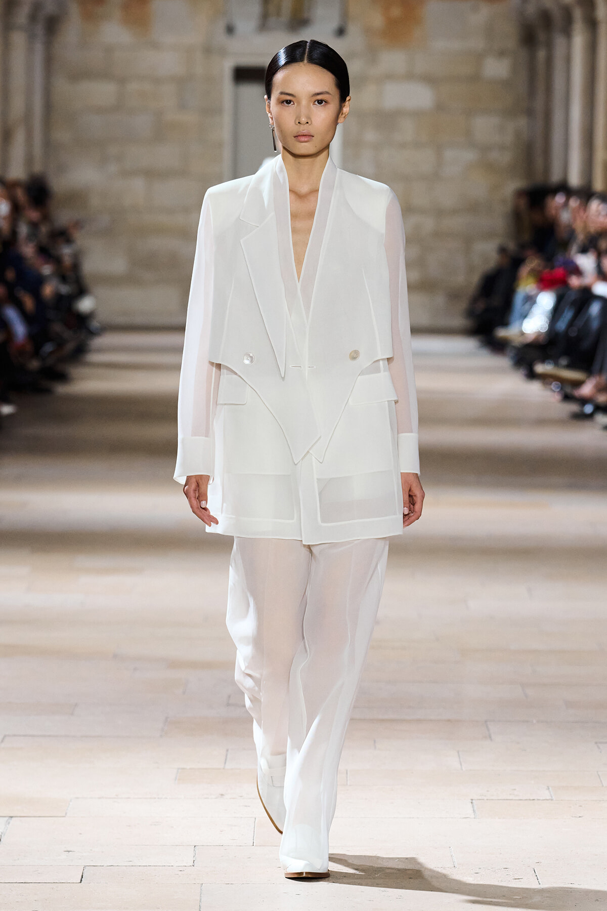 Model wearing an all-white layered blazer and wide-leg pants walks a fashion runway, sheer sleeves and structured detailing visible.