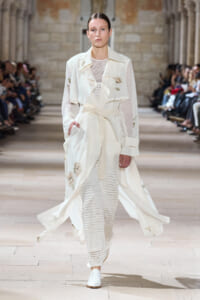 Model on a runway wearing a long white coat with subtle floral embroidery and a tied waist, over a knit white dress and white shoes.