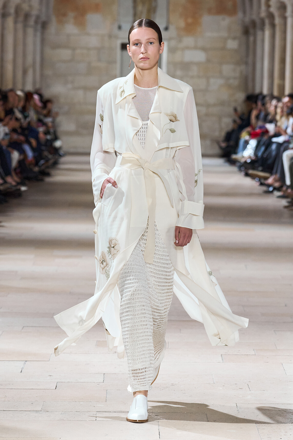 Model on a runway wearing a long white coat with subtle floral embroidery and a tied waist, over a knit white dress and white shoes.