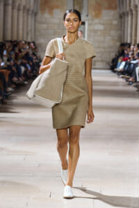 Model walks the runway in a textured beige dress, carrying a large beige tote over her shoulder.