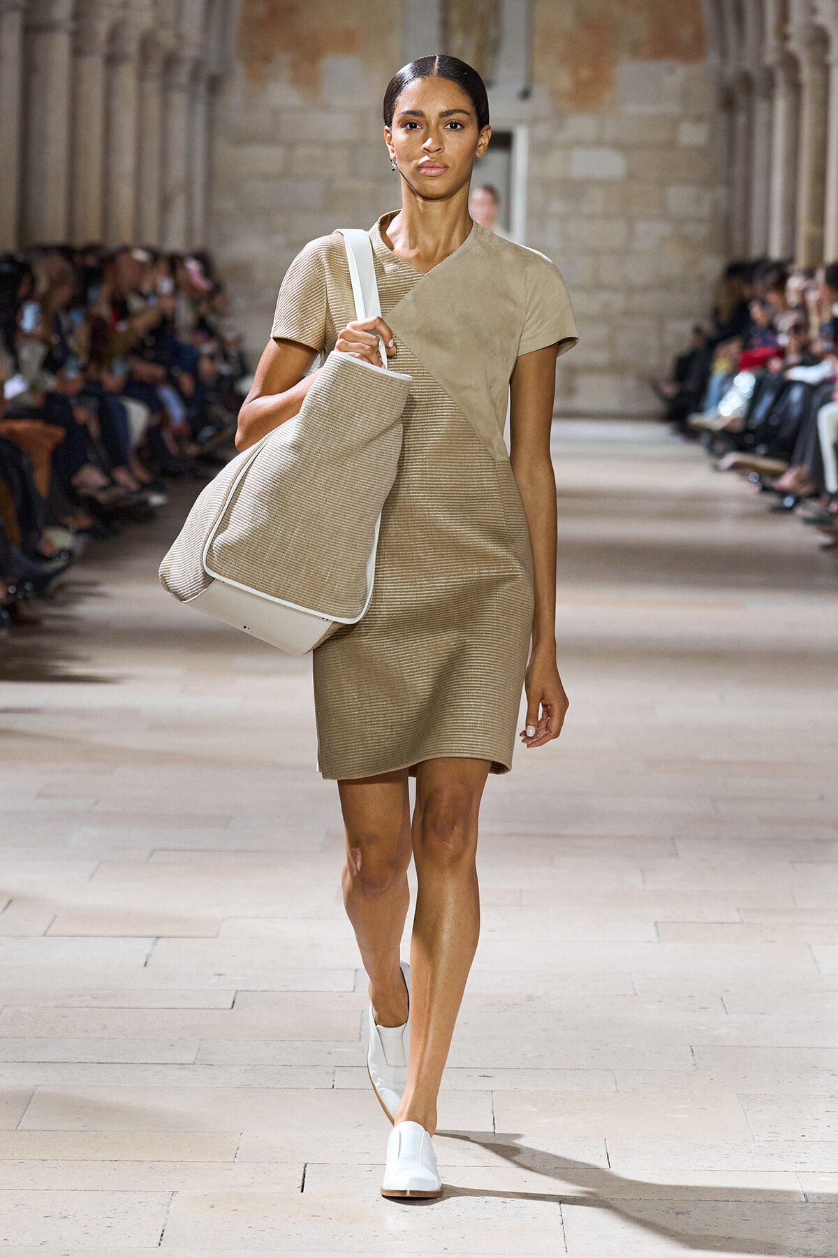 Model walks the runway in a textured beige dress, carrying a large beige tote over her shoulder.