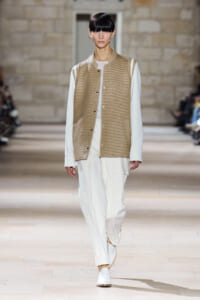 Model on a runway wearing a beige ribbed varsity jacket over a white outfit with white pants and sneakers, walking forward.