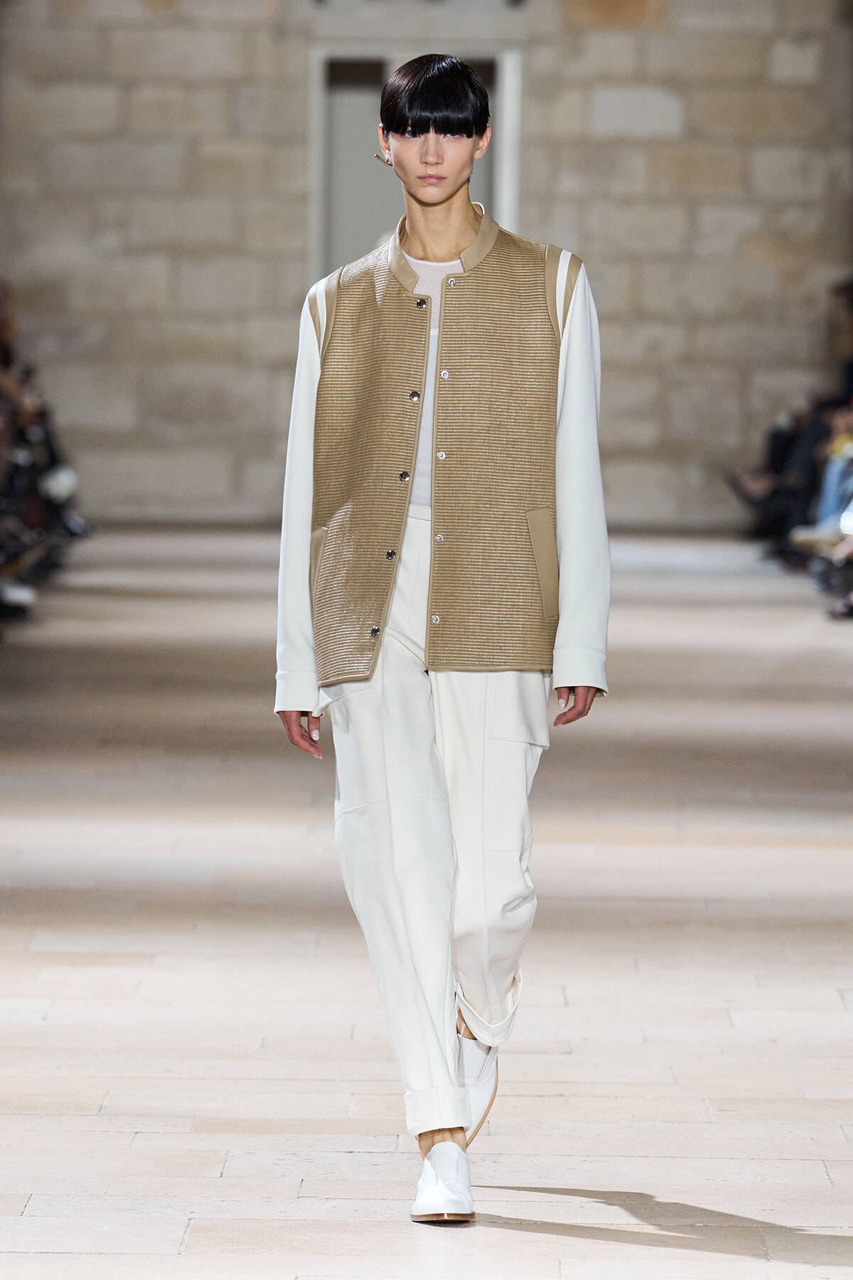 Model on a runway wearing a beige ribbed varsity jacket over a white outfit with white pants and sneakers, walking forward.