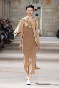 Model walks the runway in a taupe knit midi dress with a draped tan jacket and a large bow detail over one shoulder, white shoes.
