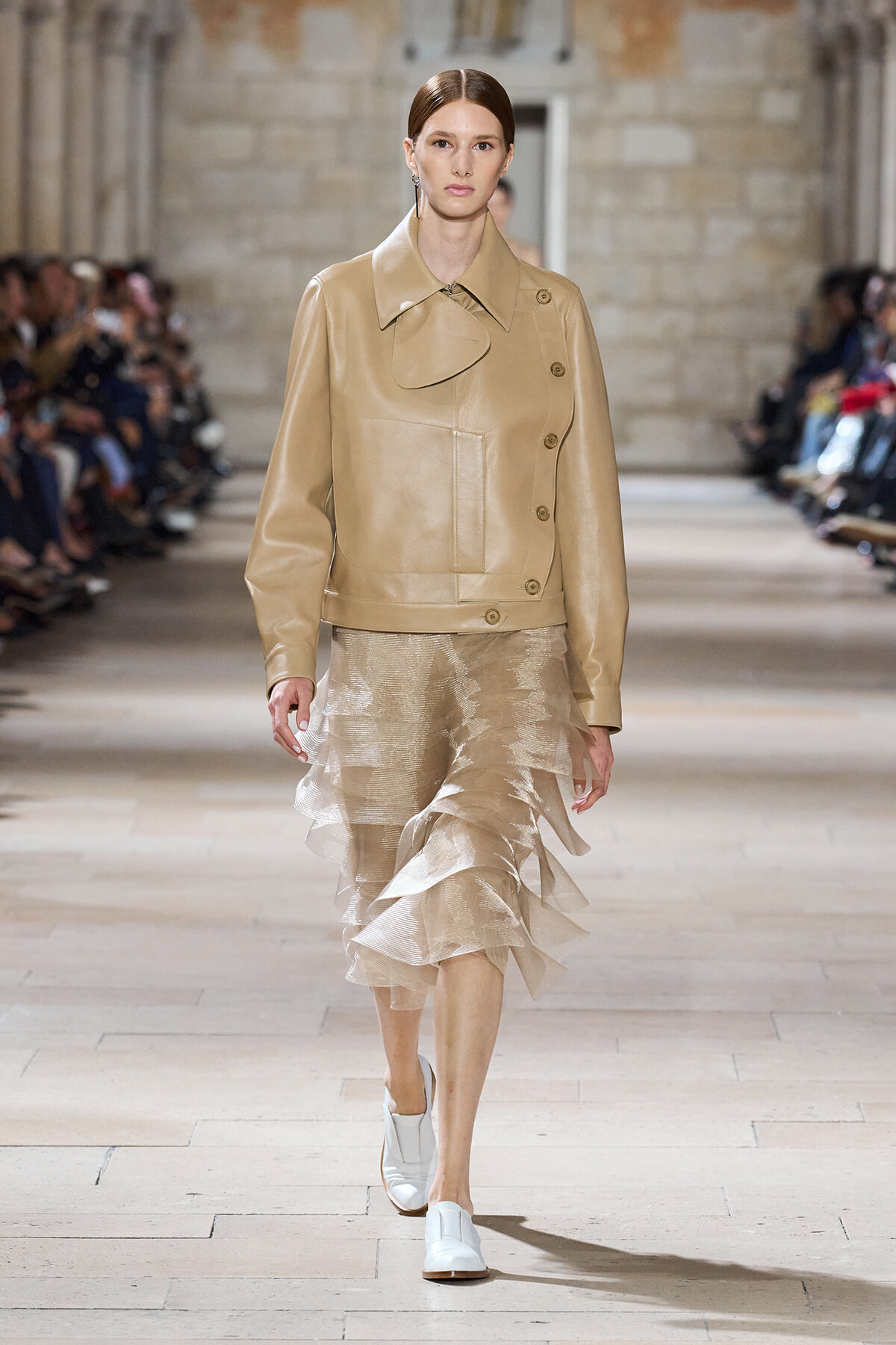 Model walks the runway in a tan leather jacket with a large front pocket and tiered beige sheer skirt at a fashion show.