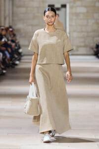 Model walks the runway in a beige ribbed knit two-piece outfit—a short-sleeve top and long skirt—carrying a beige bag with white handles.