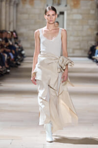 Model walking the runway in a white tank top and a layered beige draped skirt with white boots on a fashion catwalk.
