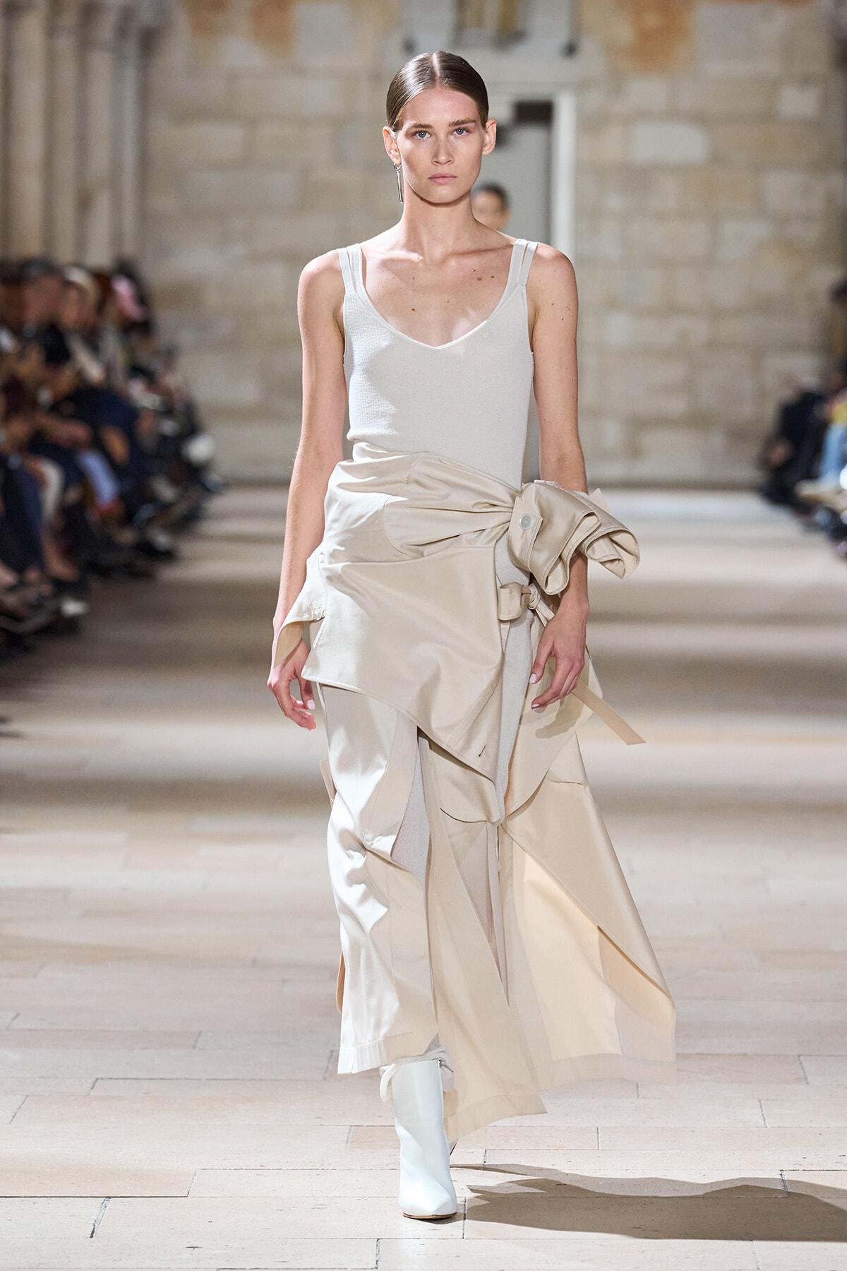 Model walking the runway in a white tank top and a layered beige draped skirt with white boots on a fashion catwalk.