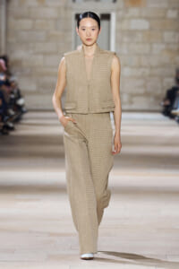 Model walks a runway wearing a coordinated beige sleeveless vest and wide-leg pants in a neutral-toned fabric, hands in pockets, desert-toned backdrop.”] ,