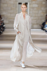 Model on a runway wearing a long ivory coat over a pale beige outfit, moving with hands in pockets.
