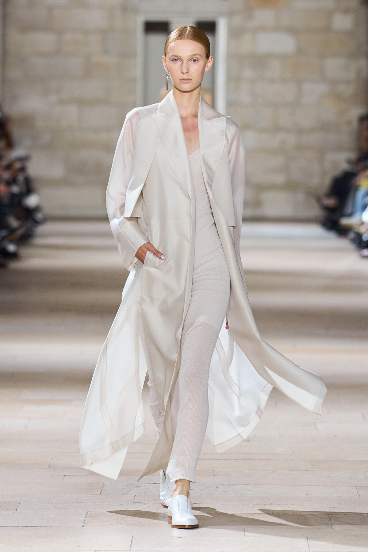 Model on a runway wearing a long ivory coat over a pale beige outfit, moving with hands in pockets.