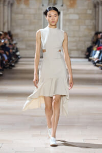 Model on a runway wearing a sleeveless ivory high-neck dress with waist cutouts and a high-low flared hem.