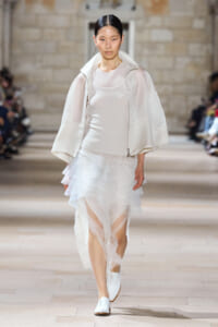 Model on a runway wearing an all-white outfit: sheer tiered skirt, white top, and a light jacket with white shoes.