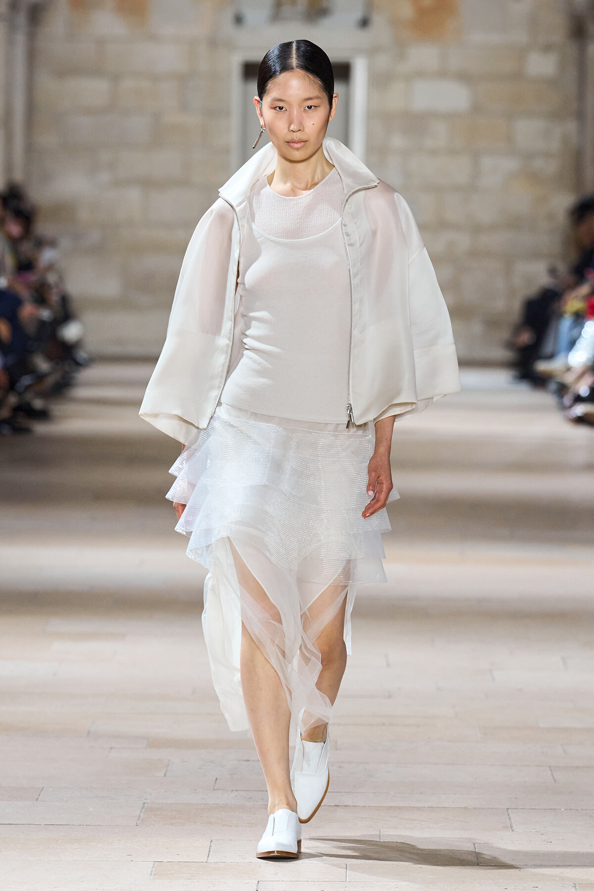 Model on a runway wearing an all-white outfit: sheer tiered skirt, white top, and a light jacket with white shoes.