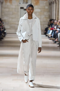 Model walking a fashion runway in an all-white layered trench coat over a knit dress, paired with white loafers.