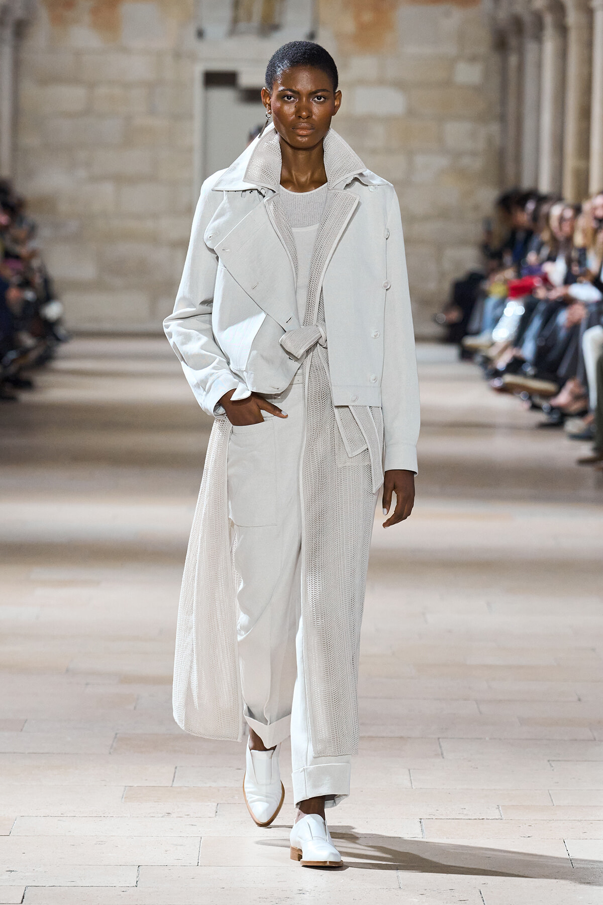 Model walking a fashion runway in an all-white layered trench coat over a knit dress, paired with white loafers.