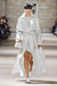 Model walks a runway in a pale trench coat with a tied belt and white accessories, mid-stride on a catwalk.