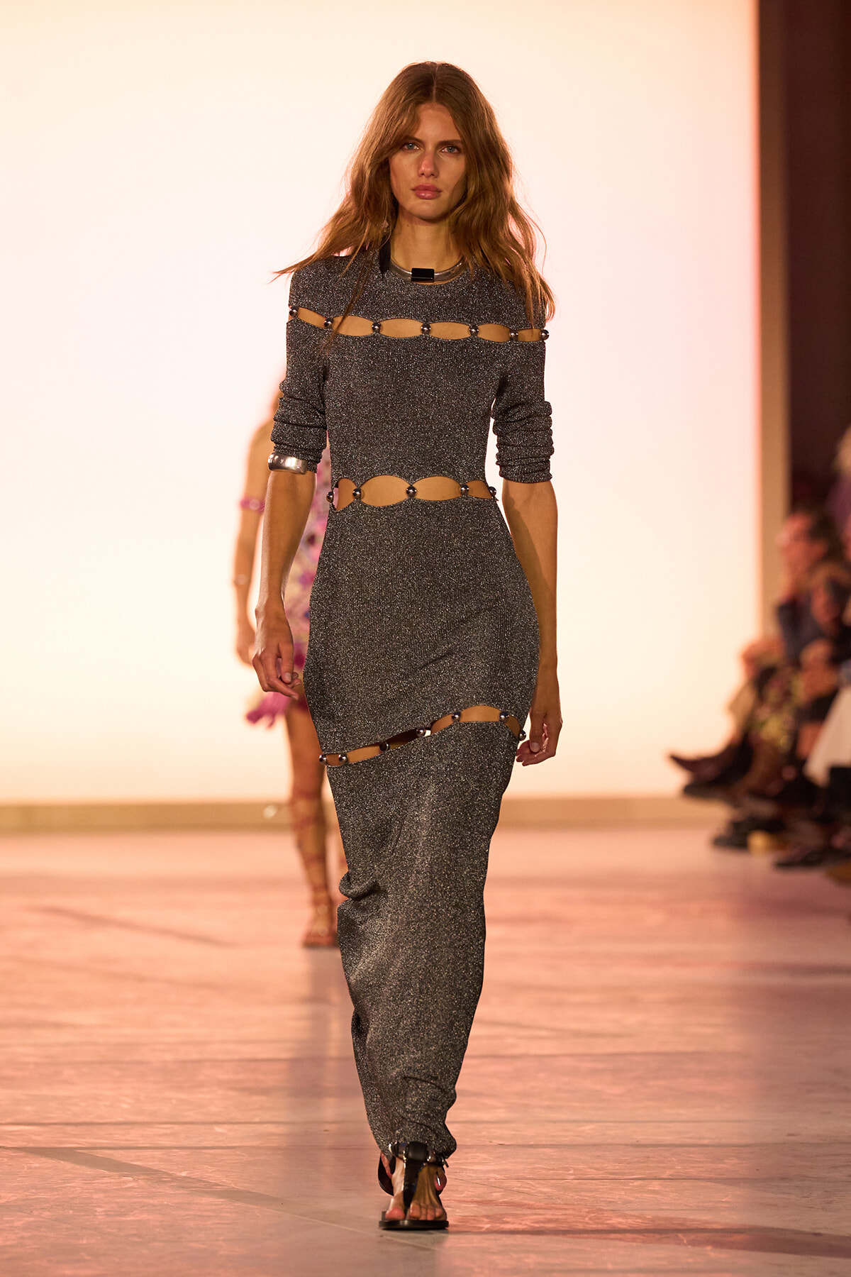 Model walks a runway wearing a glittery gray dress with cut-out belt details around the waist and shoulders, on a fashion show catwalk.