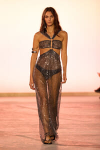 Model walking the runway in a metallic, sequined halter top with cutouts and sheer glitter pants; black underwear visible, bold accessories.