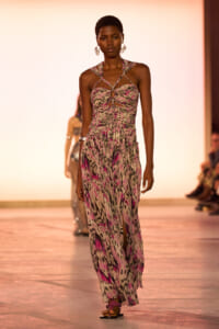 Model walks a fashion runway in a pink‑purple abstract print halter dress with cutouts and crisscross straps, pink sandals.